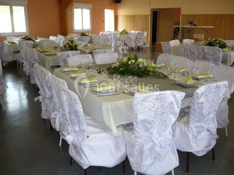 Salle décorée pour un événement avec des tables dressées, nappes blanches, chaises couvertes et arrangements floraux.