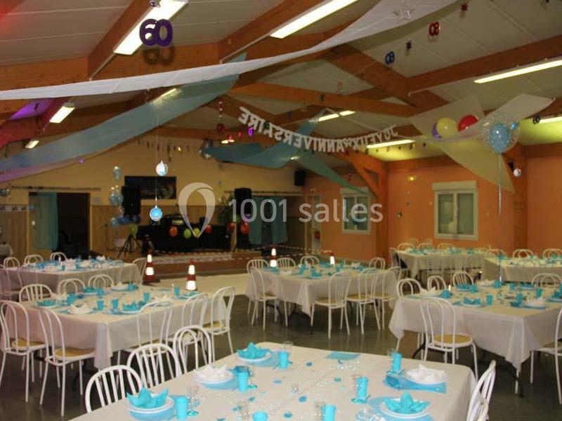 Salle décorée pour une fête, avec des tables dressées, des ballons et des guirlandes suspendues au plafond.