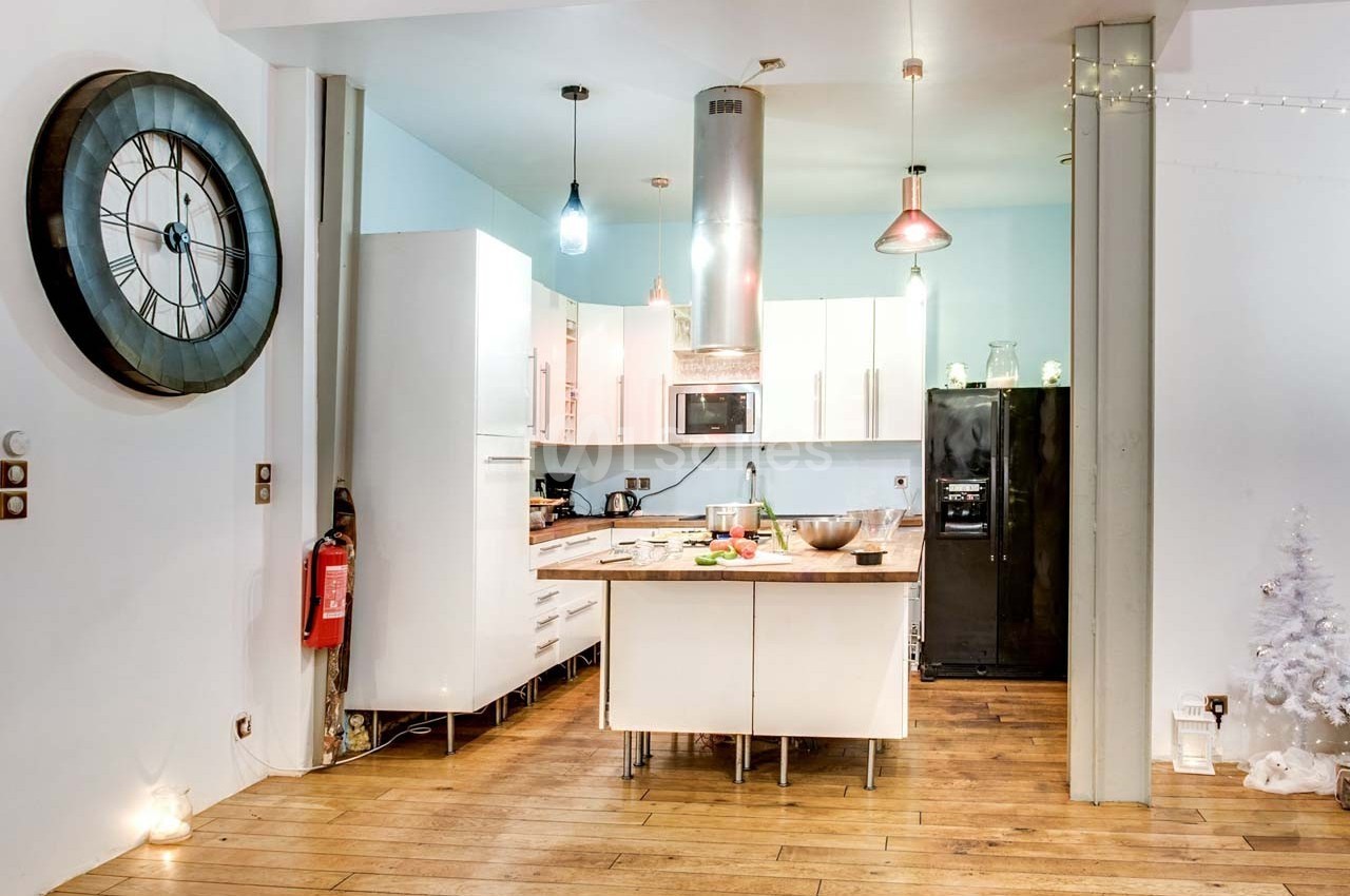 Cuisine moderne avec îlot central, sol en bois, murs clairs, horloge murale et décorations lumineuses.