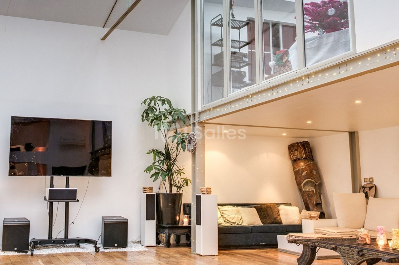 Salon moderne avec parquet, canapé, plantes, télévision murale et mezzanine vitrée décorée de lumières.