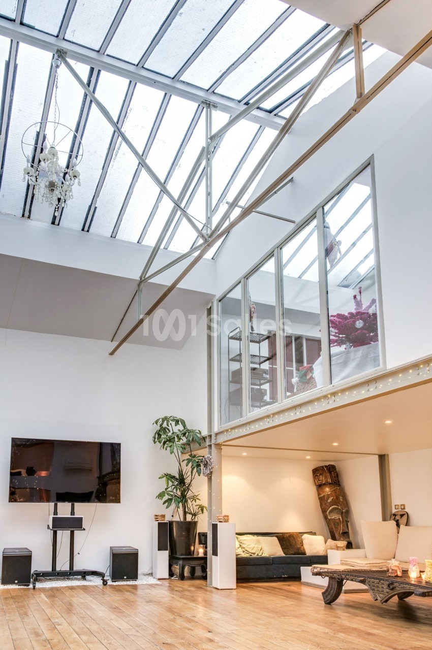 Salon lumineux avec parquet, grande verrière, canapé, plantes, télévision murale et mezzanine.