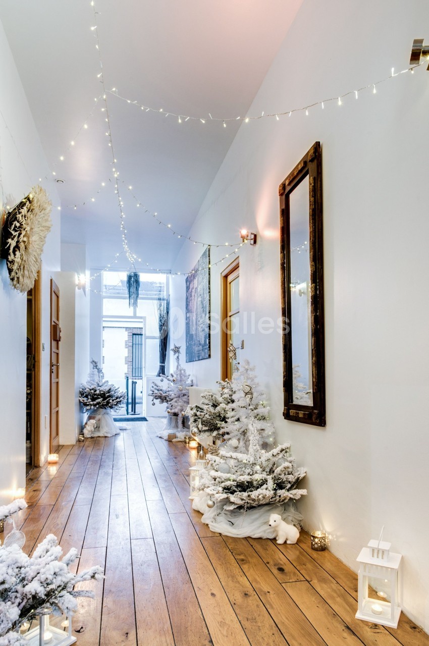Couloir décoré pour Noël avec guirlandes lumineuses, sapins enneigés, lanternes blanches et sol en bois clair.