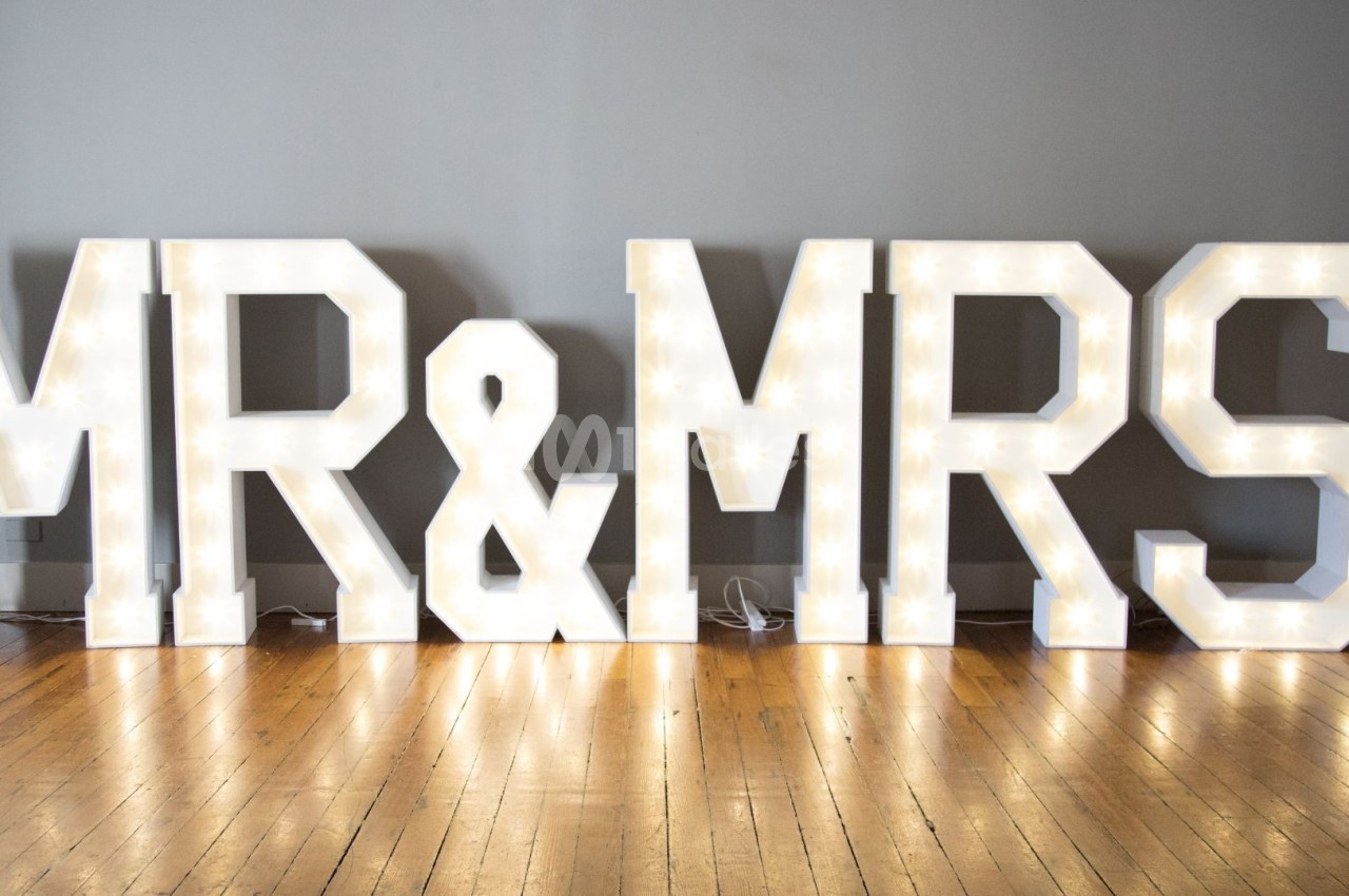 Lettres lumineuses formant ’MR & MRS’ posées sur un sol en bois devant un mur gris.