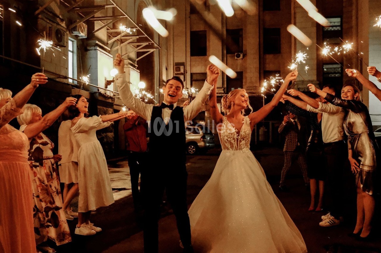 Un couple en tenue de mariage entouré d'invités tenant des cierges magiques allumés dans une cour en soirée.