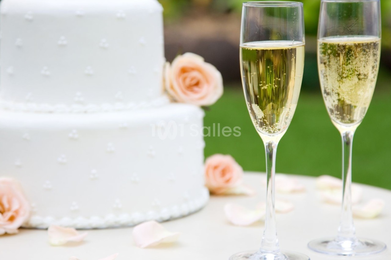 Gâteau de mariage blanc décoré de perles et deux coupes de champagne sur une table avec des pétales de rose.
