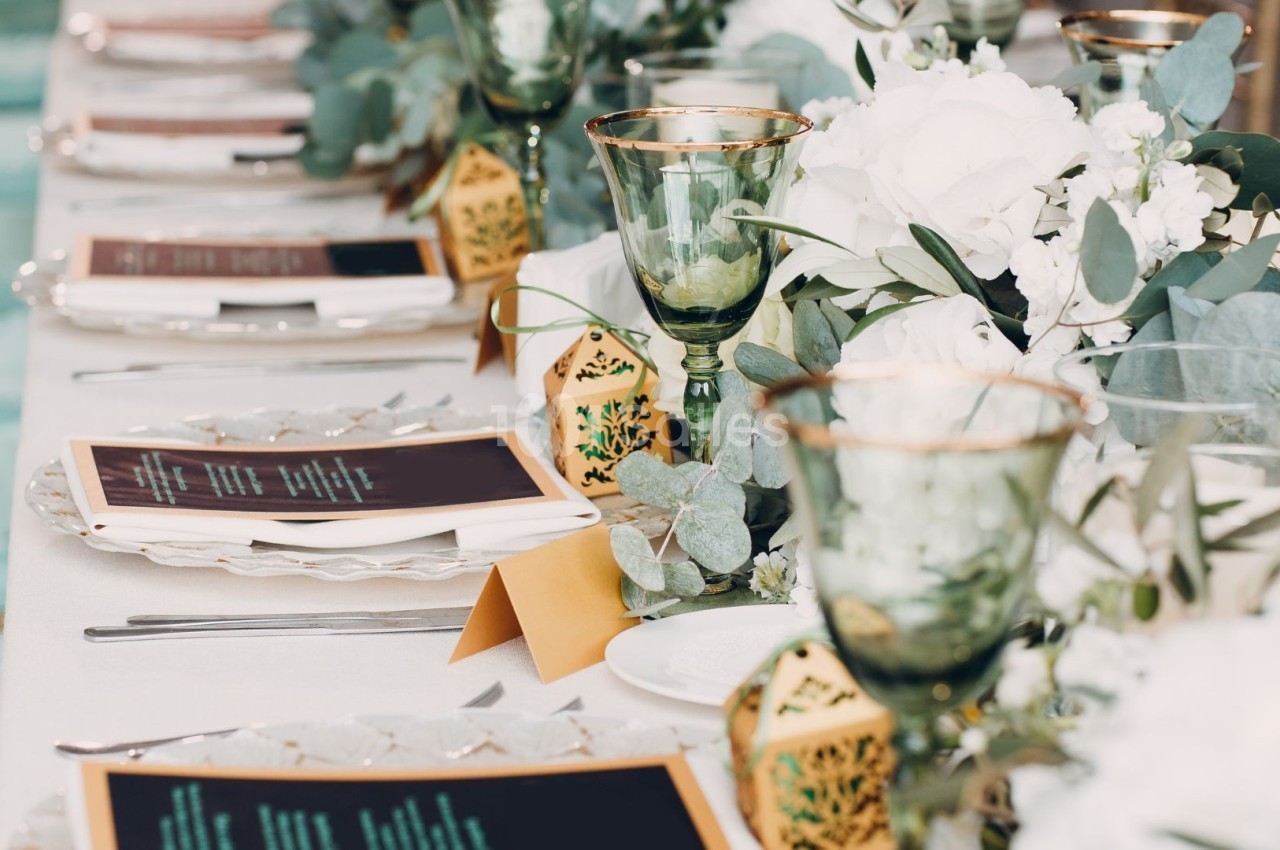 Table élégamment dressée avec des menus, des verres dorés, des fleurs blanches et des décorations végétales.