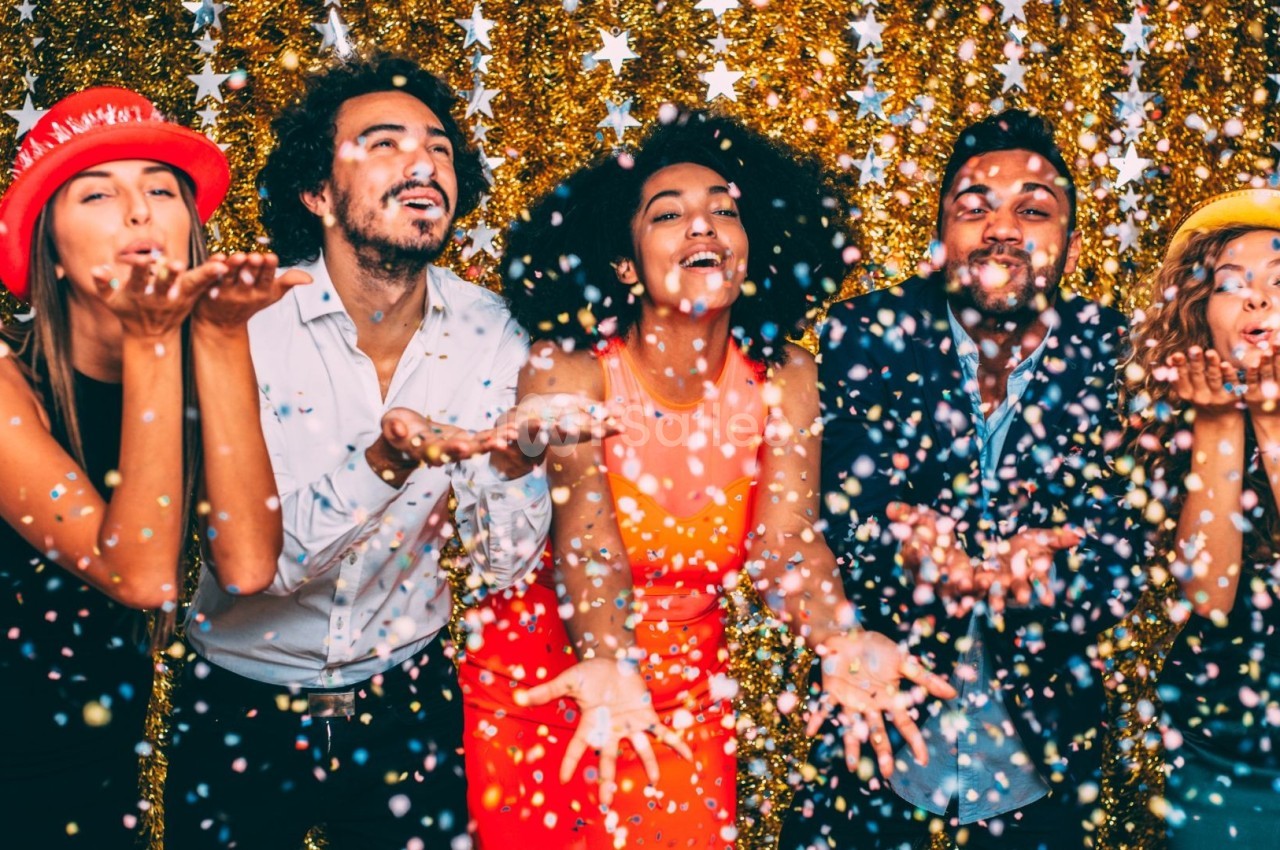 Des personnes souriantes soufflent des confettis colorés devant un fond doré décoré d'étoiles.