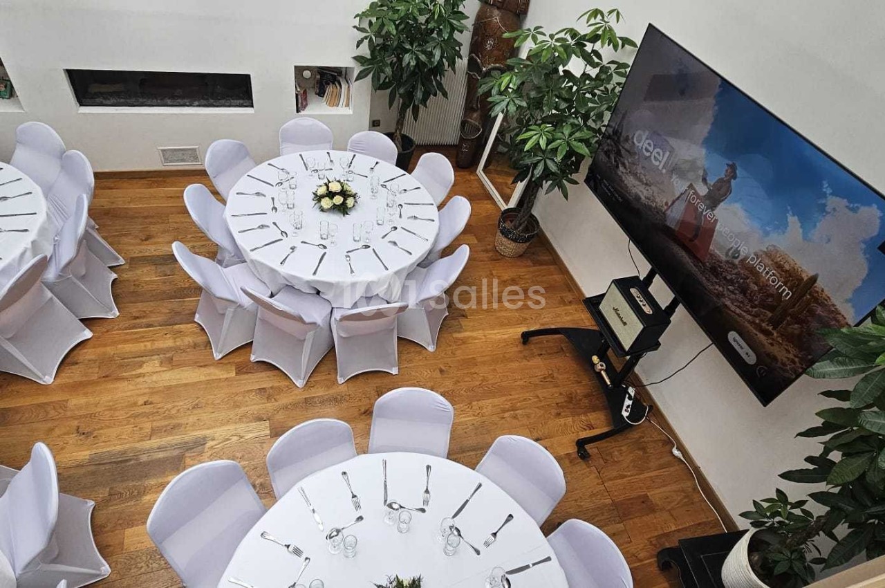 Salle lumineuse avec tables rondes dressées, nappes blanches, bouquets de fleurs et écran mural affichant une image.