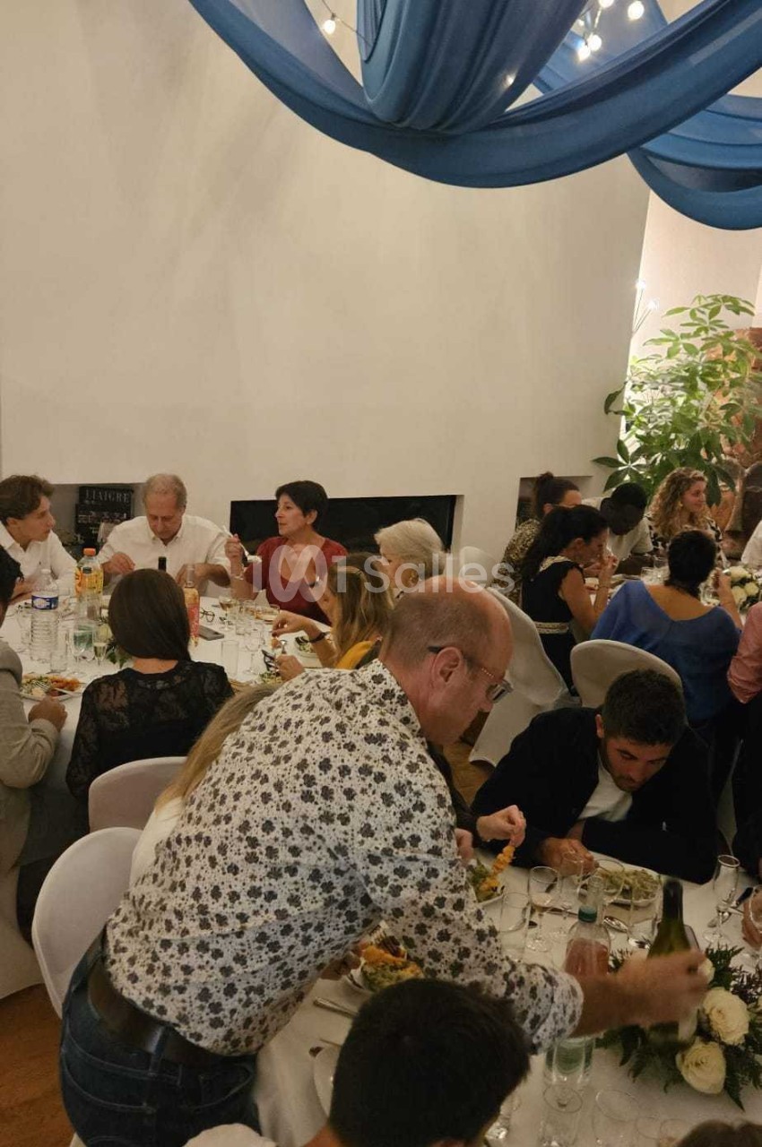 Des convives assis autour de tables décorées, partageant un repas dans une salle ornée de drapés bleus et de guirlandes…