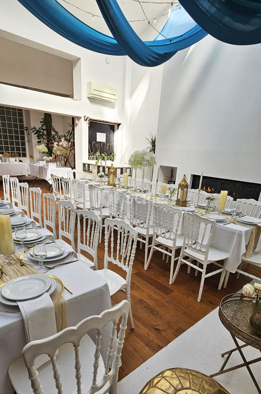 Salle de réception lumineuse avec tables dressées, chaises blanches, décorations dorées et voilages bleus suspendus.