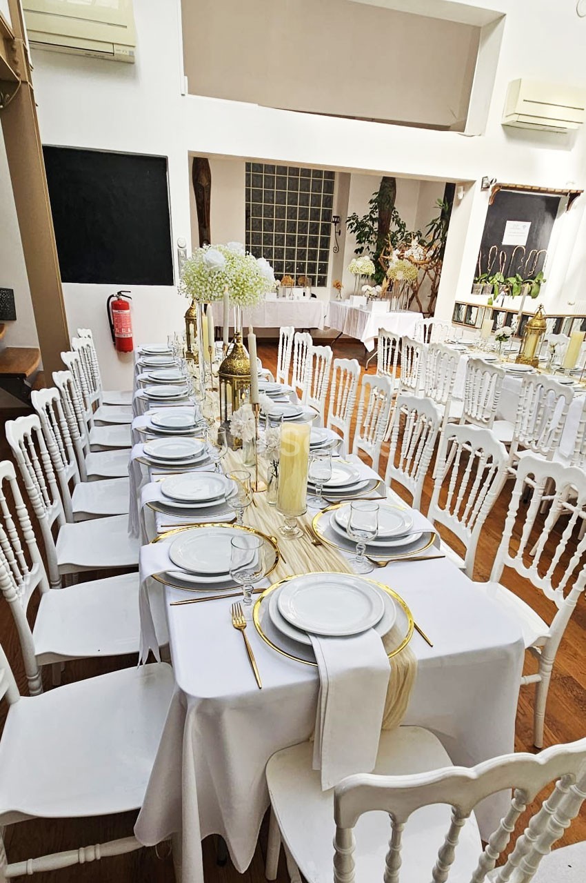 Salle décorée pour un événement avec tables alignées, nappes blanches, vaisselle élégante et chaises blanches.