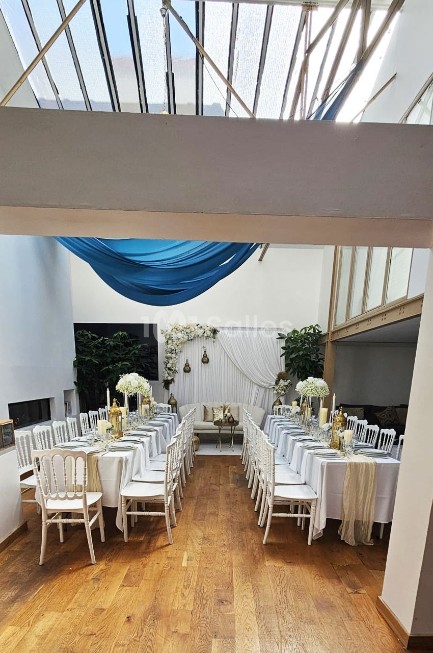 Salle décorée pour un événement, avec tables alignées, nappes blanches, chaises élégantes et ornements floraux.