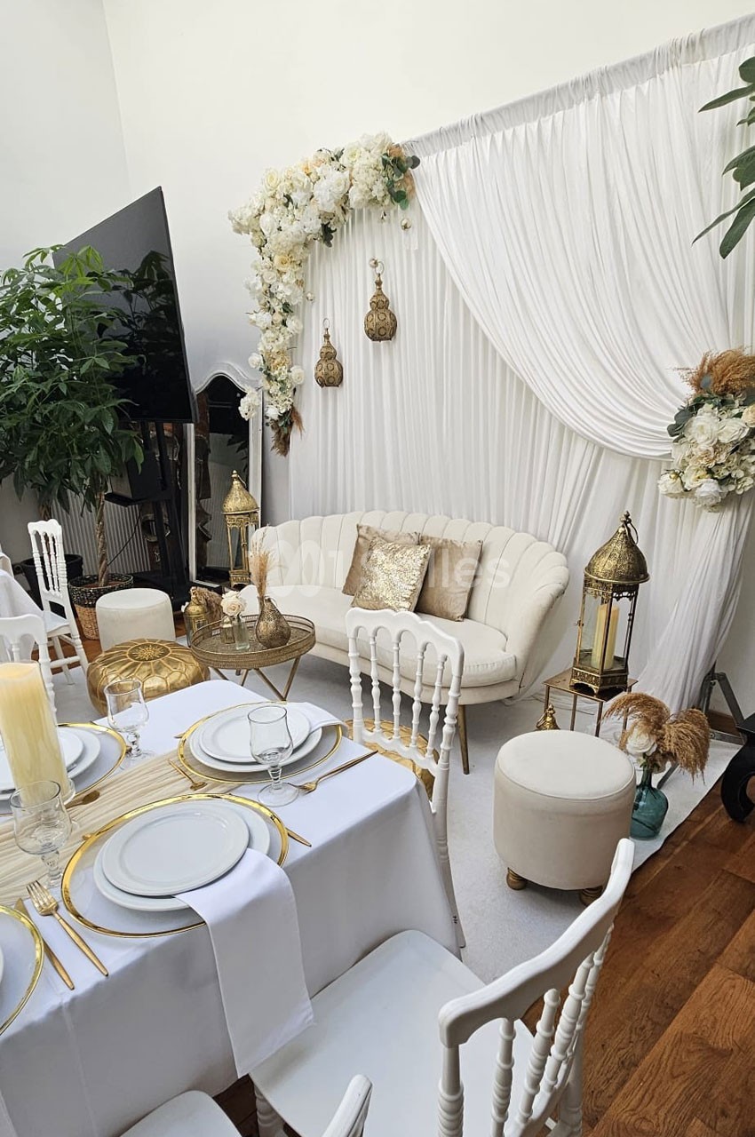 Salon décoré avec un canapé crème, des lanternes dorées, des fleurs blanches et une table dressée élégamment.