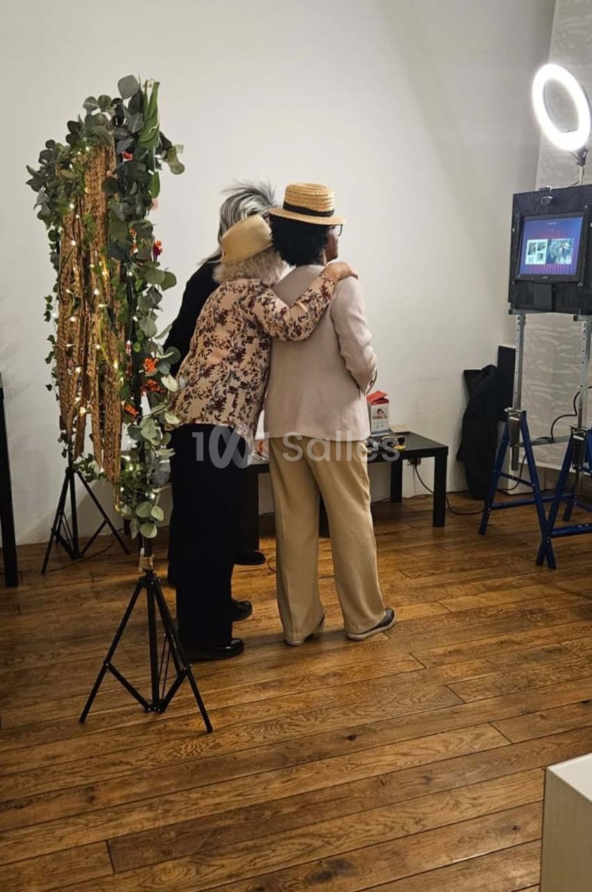 Deux personnes de dos, portant des chapeaux, posent devant un décor floral et un éclairage de studio.