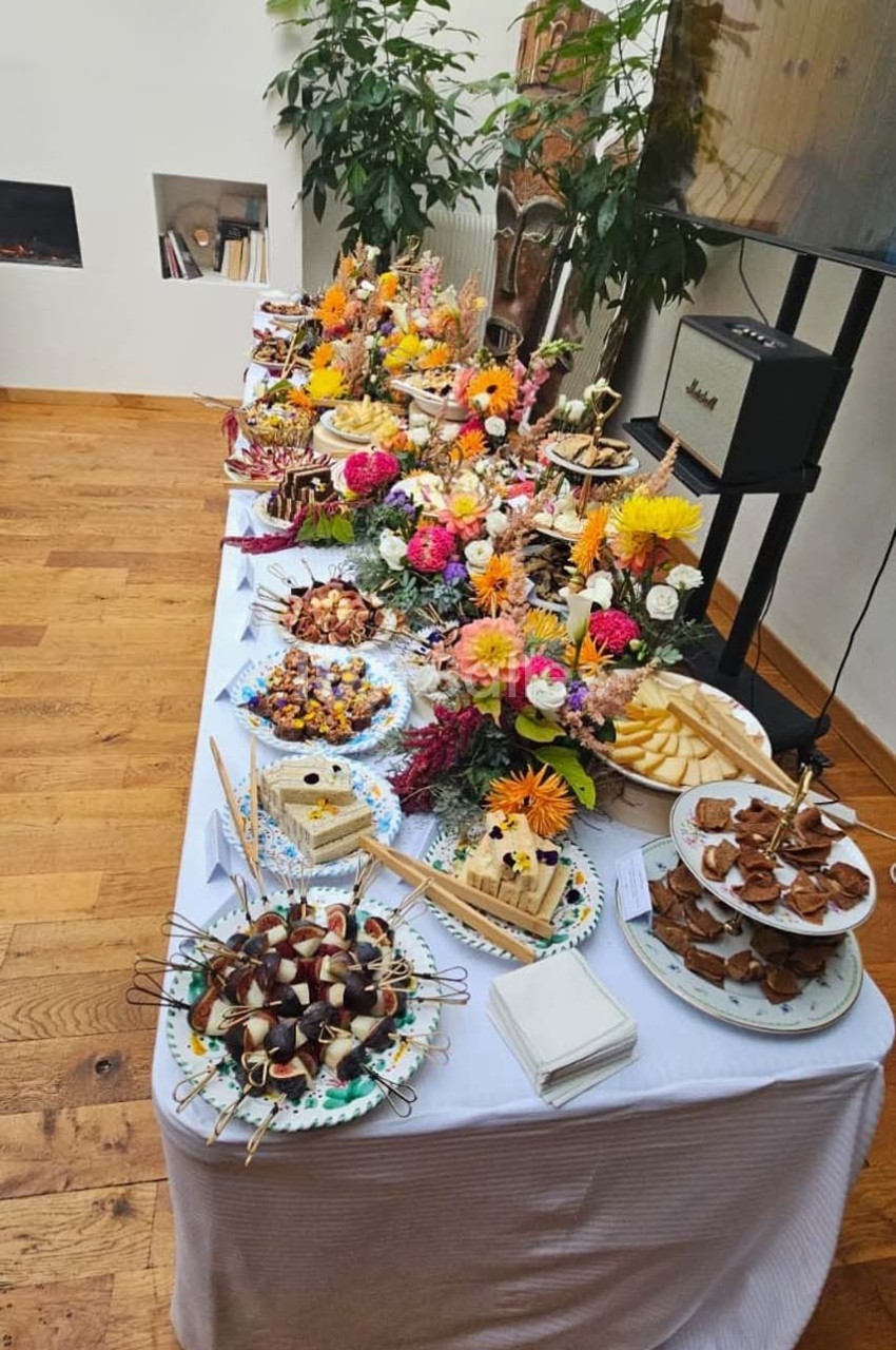 Table garnie de plats variés, desserts et fleurs colorées, disposée dans une pièce lumineuse avec parquet en bois.