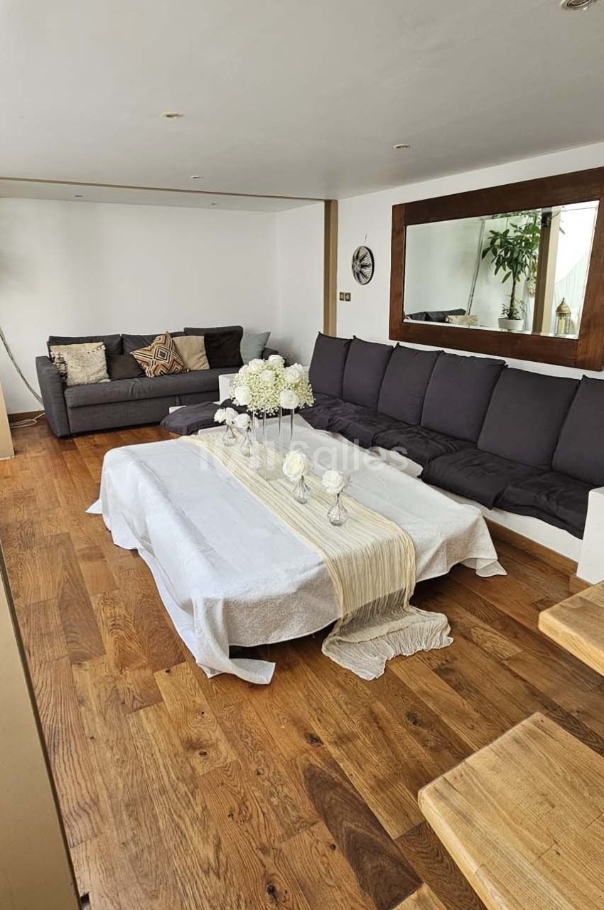 Salon avec parquet, table basse décorée de fleurs blanches, canapés gris et grand miroir mural.