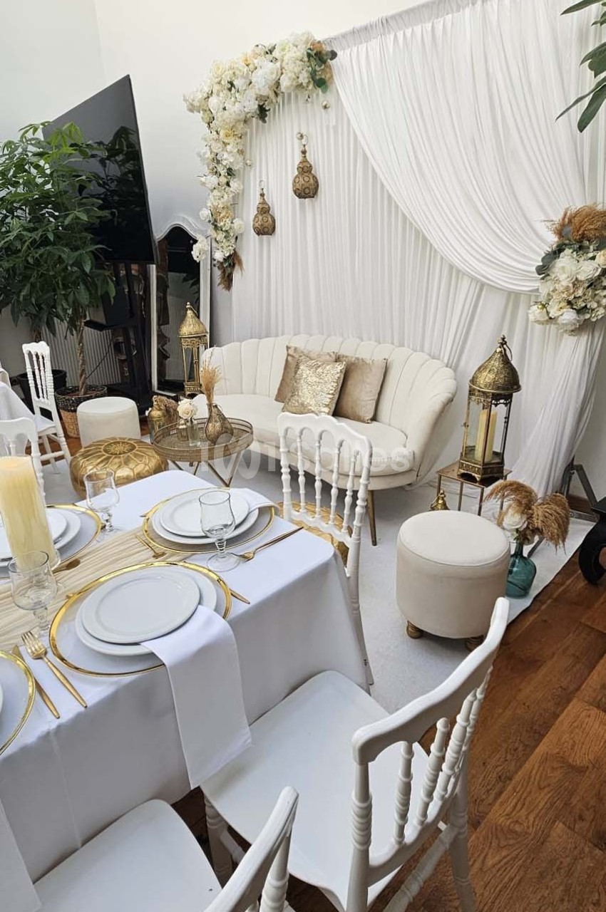 Salon décoré avec un canapé crème, rideaux blancs, fleurs, lanternes dorées et table dressée avec vaisselle élégante.