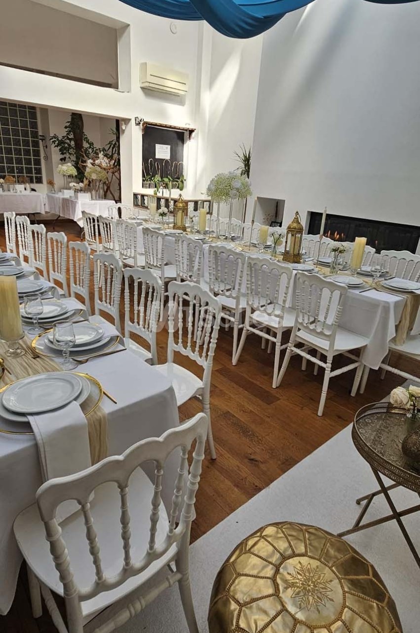 Salle lumineuse décorée pour un événement, avec tables dressées, chaises blanches et éléments dorés.