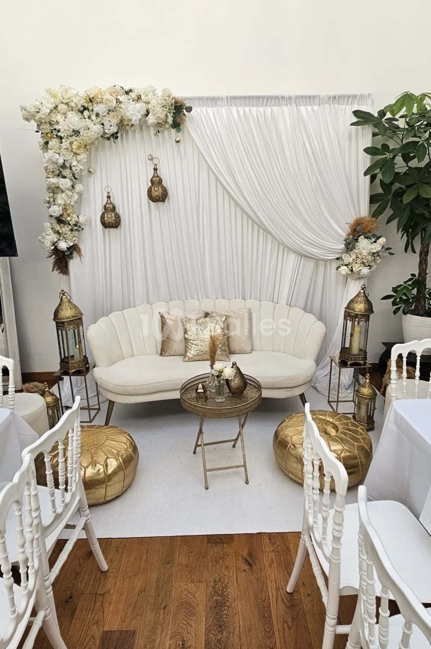 Décor élégant avec canapé blanc, coussins dorés, lanternes, poufs et rideaux blancs ornés de fleurs.