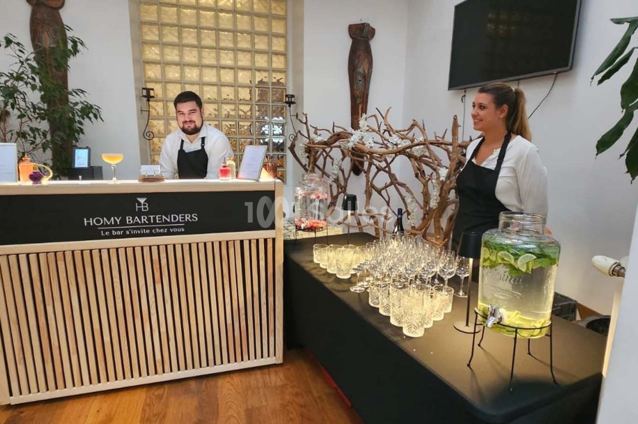 Deux personnes derrière un comptoir de bar avec des verres, des boissons et une fontaine d'eau aromatisée.