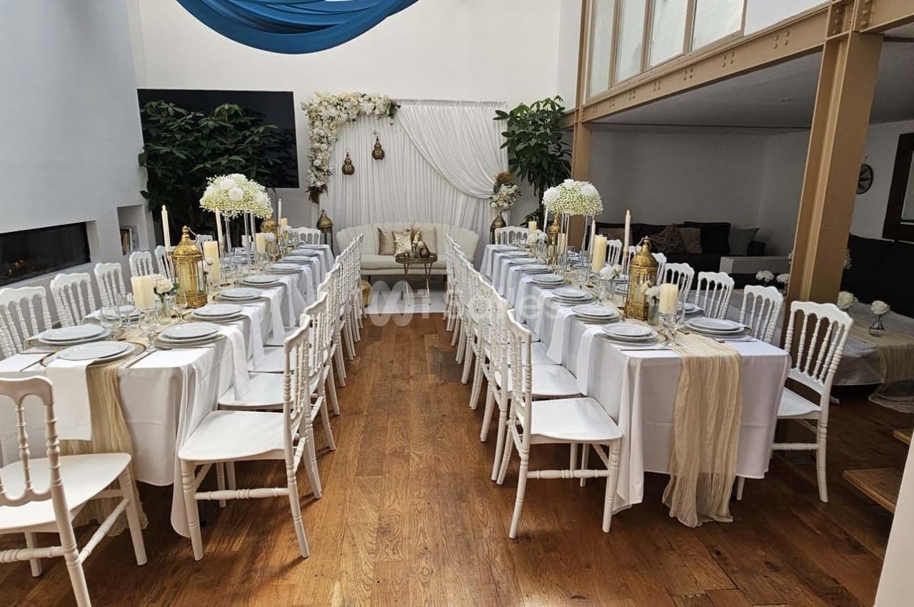 Salle décorée pour un événement avec deux longues tables dressées, nappes blanches, bougies et fleurs blanches.