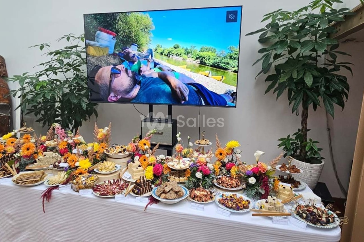 Table garnie de plats variés et décorée de fleurs colorées, avec un écran affichant une scène extérieure en arrière-plan.