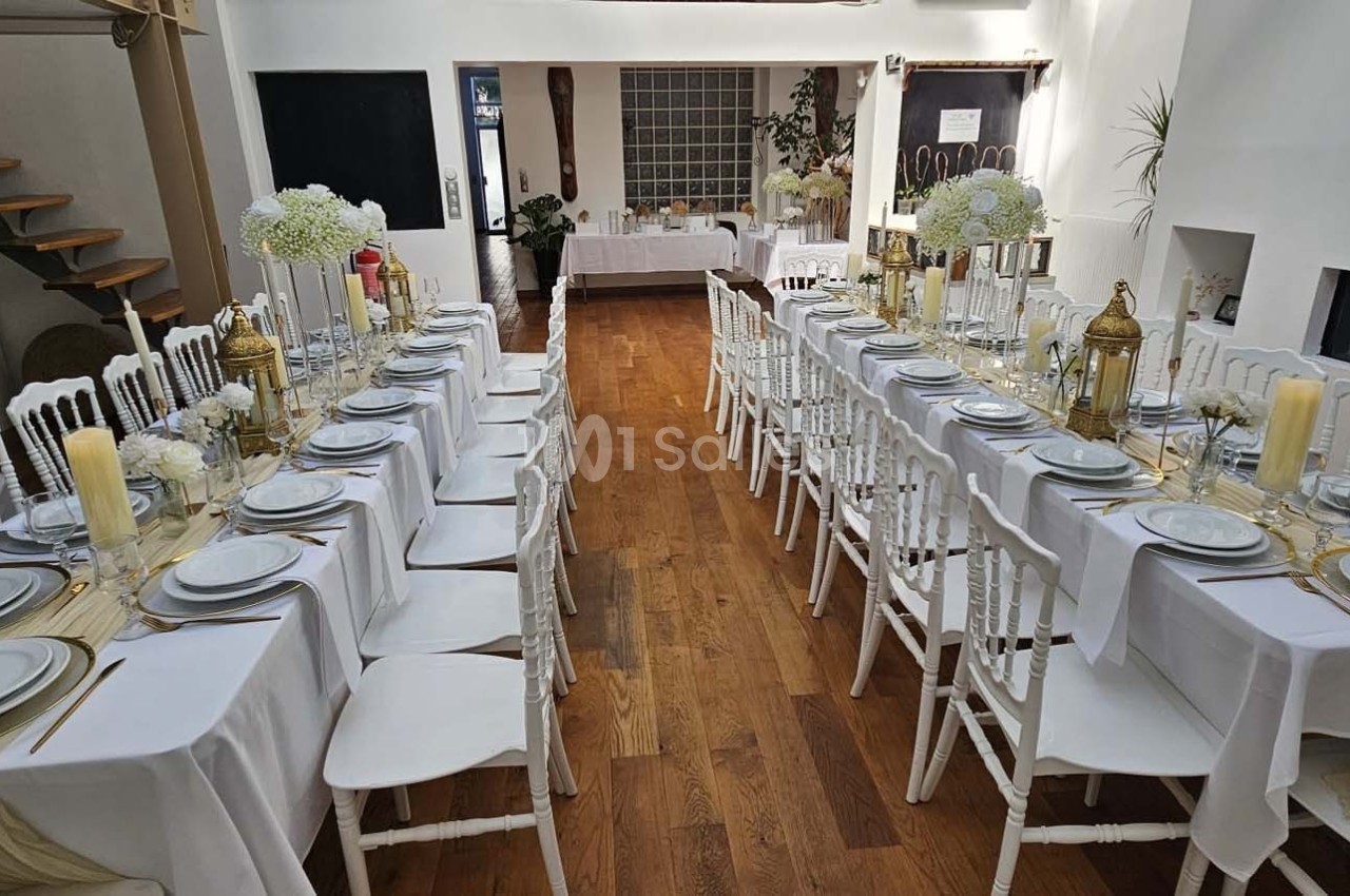 Salle décorée pour un repas, avec deux longues tables dressées, nappes blanches, vaisselle élégante et lanternes dorées.