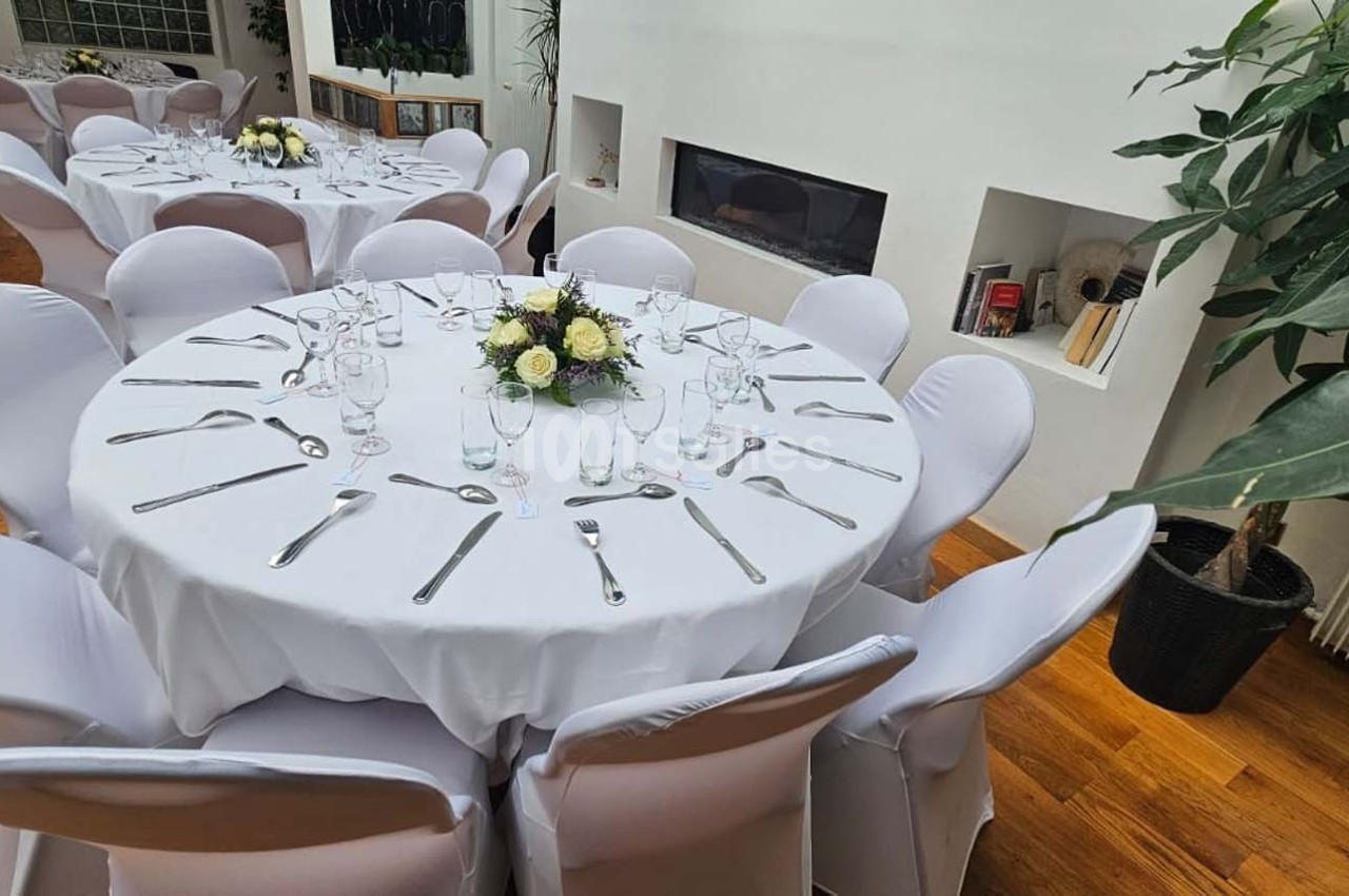 Tables rondes dressées avec nappes blanches, couverts et centres de table fleuris, dans une salle lumineuse avec plantes.