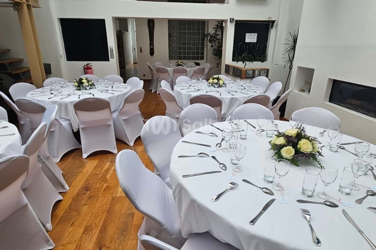 Salle lumineuse avec tables rondes dressées de nappes blanches, couverts et centres de table fleuris.