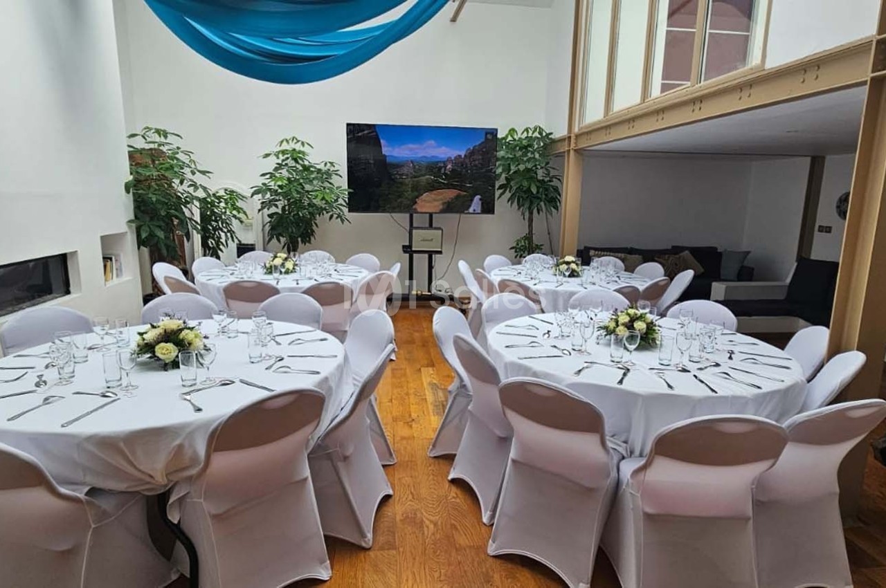 Salle lumineuse avec tables rondes dressées, nappes blanches, décor floral et écran affichant un paysage.