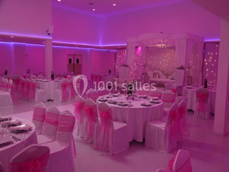 Salle de réception décorée en rose et blanc avec des tables dressées et une estrade ornée de rideaux lumineux.