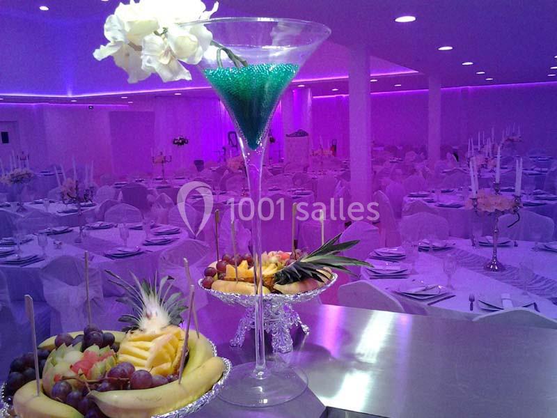 Salle de réception éclairée en violet avec tables dressées et coupes de fruits décorées au premier plan.