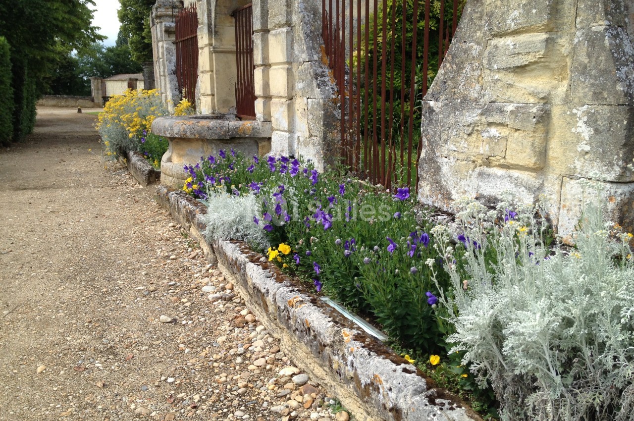 Parterre de fleurs colorées bordant un vieux mur en pierre avec des grilles métalliques et un chemin de gravier.