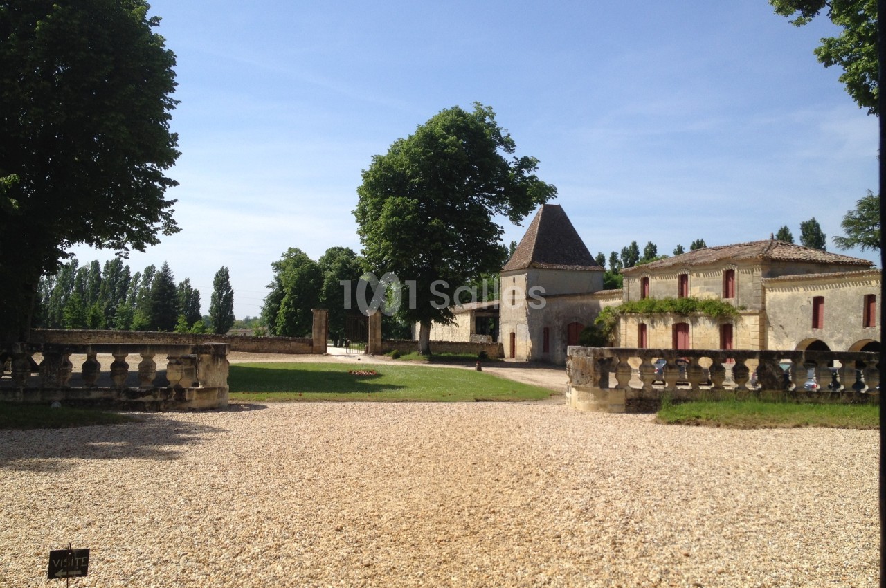 Cour d’un domaine avec bâtiments en pierre, allée gravillonnée, balustrades et arbres sous un ciel dégagé.