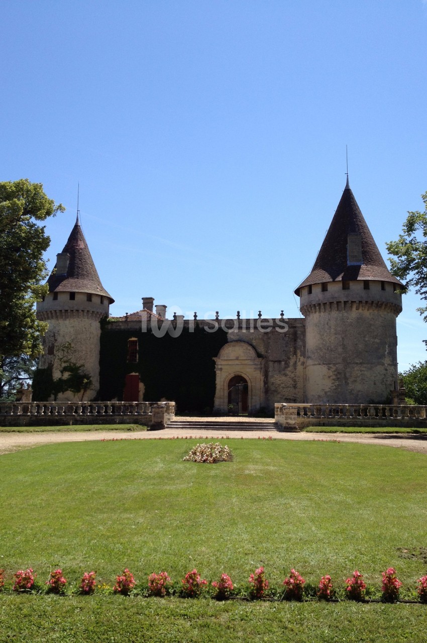Château en pierre avec deux tours, entouré de pelouse et de fleurs, sous un ciel bleu dégagé.