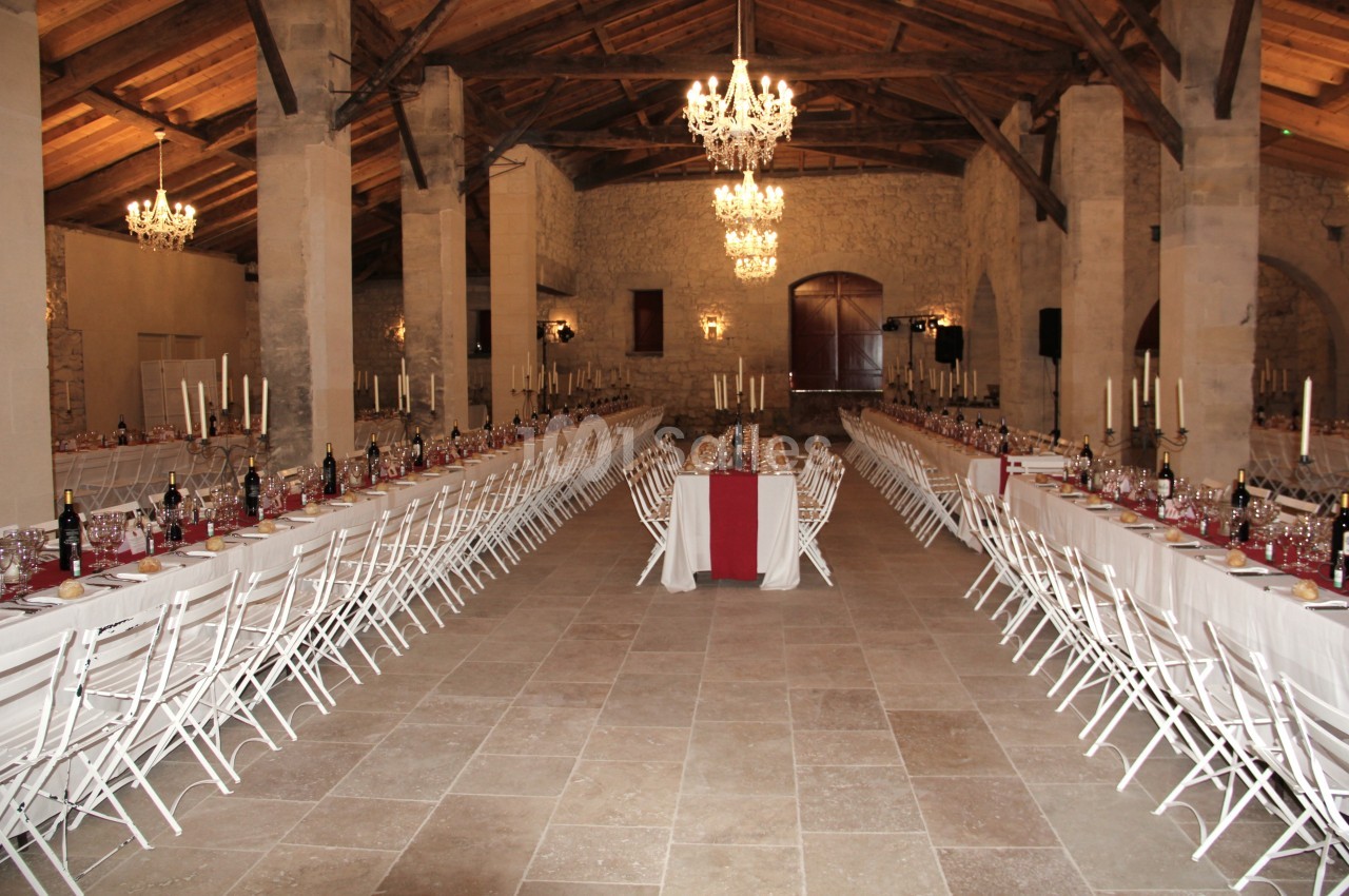 Grande salle de réception en pierre avec tables longues dressées, chaises blanches et chandeliers suspendus.