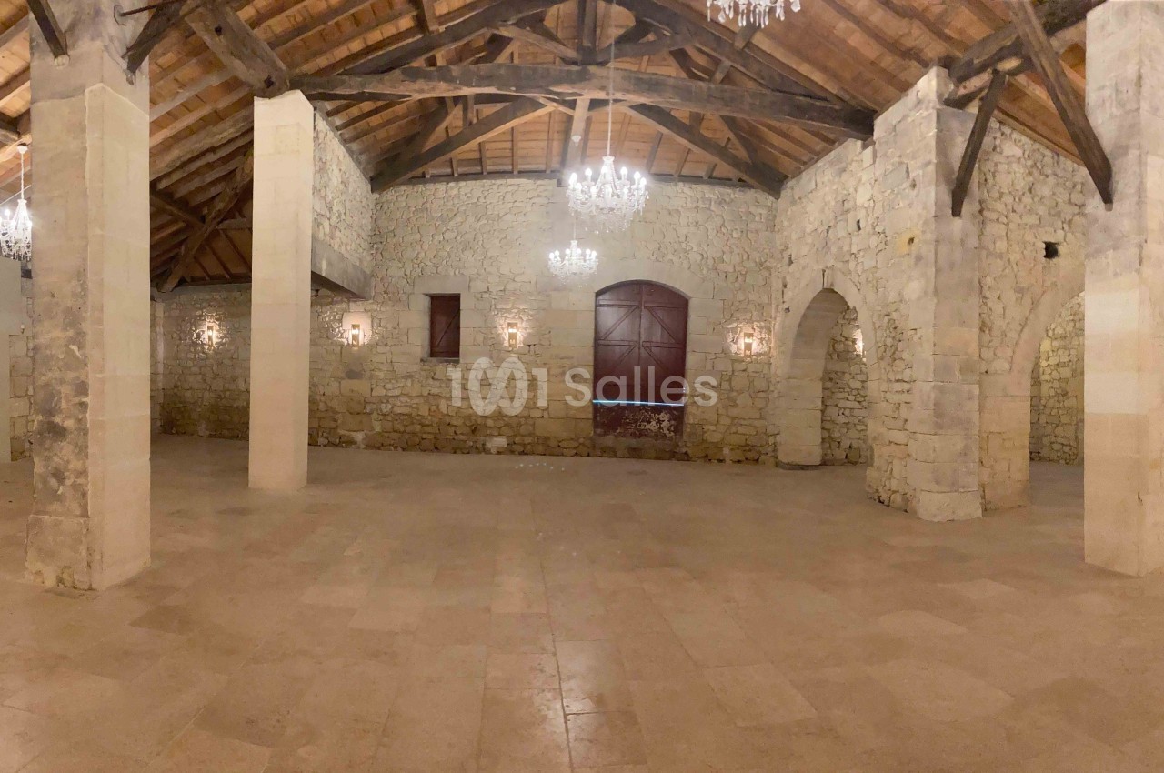 Salle en pierre avec poutres apparentes, sol en pierre claire, lustres suspendus et grandes ouvertures en bois.