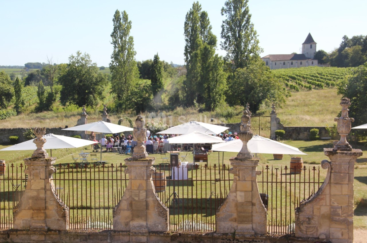 Vue d'un jardin avec des tables et parasols, entouré de vignes, et une église en arrière-plan.