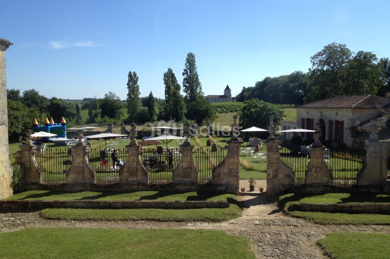 Vue d'un jardin avec pelouse, tables, parasols et une structure gonflable, entouré de bâtiments anciens et d'arbres.