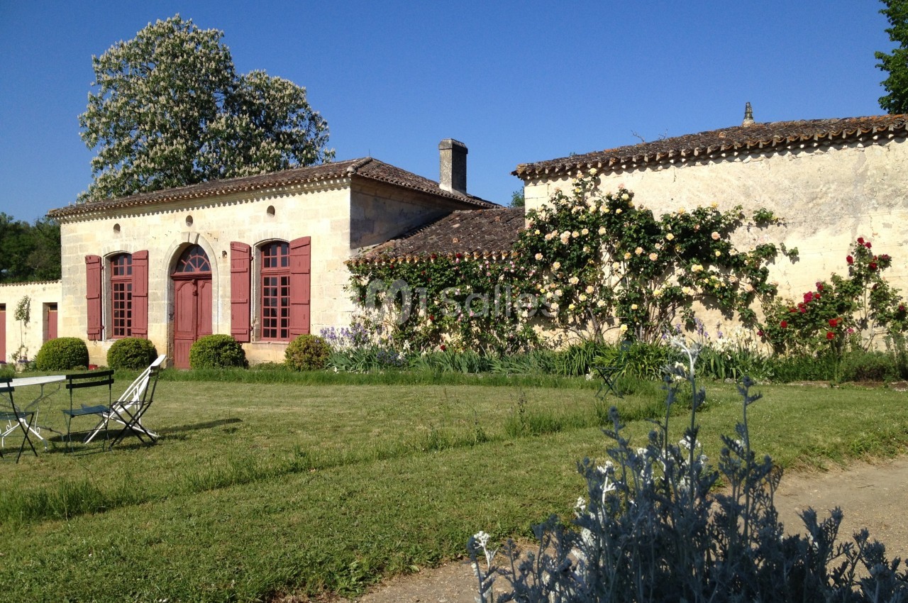 Façade d'une maison en pierre avec volets rouges, entourée de verdure et de fleurs, sous un ciel bleu.
