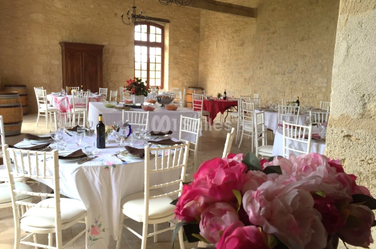Salle en pierre avec tables rondes dressées pour un repas, décorée de fleurs et éclairée par une grande fenêtre.