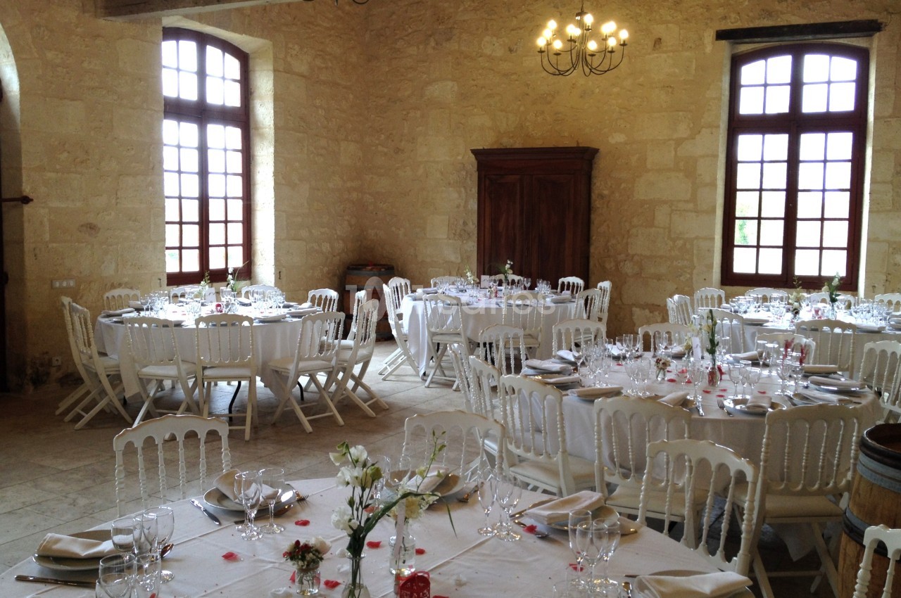 Salle de réception lumineuse avec tables rondes dressées, chaises blanches et décorations florales.
