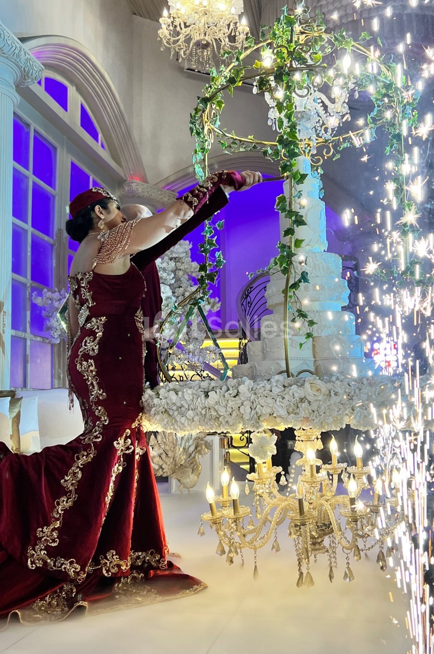 Une femme en robe rouge ornée de dorures ajuste un grand gâteau décoré de fleurs, entourée de chandeliers et d'étincelles.