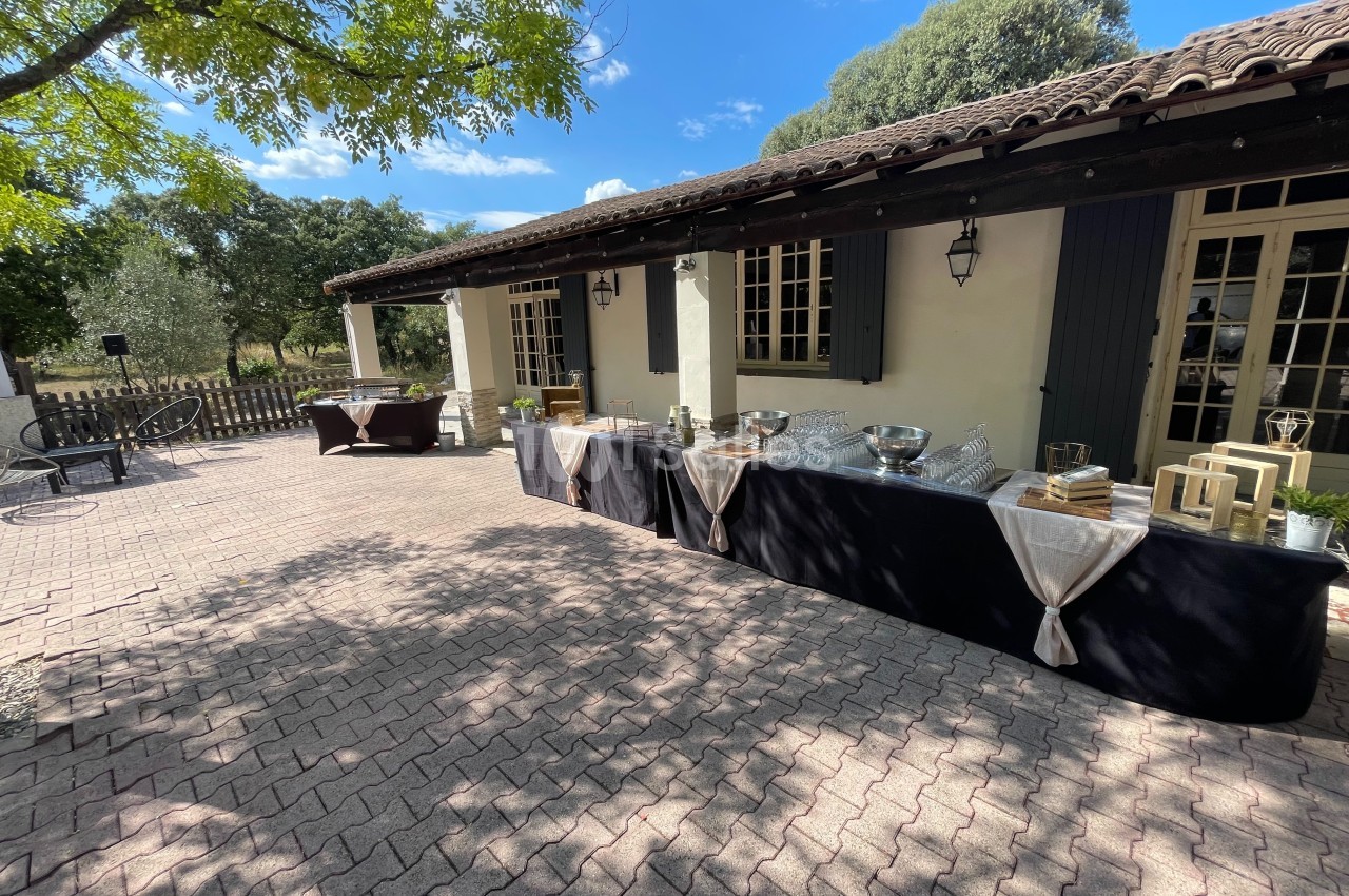 Tables de buffet dressées en extérieur sur une terrasse pavée, devant un bâtiment aux volets bleus.