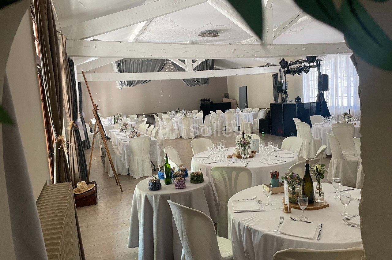 Salle de réception lumineuse avec tables rondes dressées, décorations florales et équipement de sonorisation.