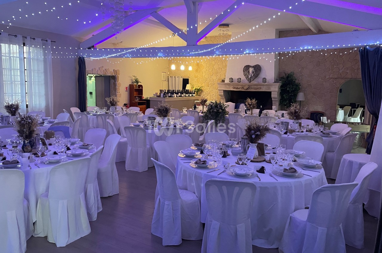 Salle de réception décorée avec des tables rondes dressées, nappes blanches et guirlandes lumineuses au plafond.