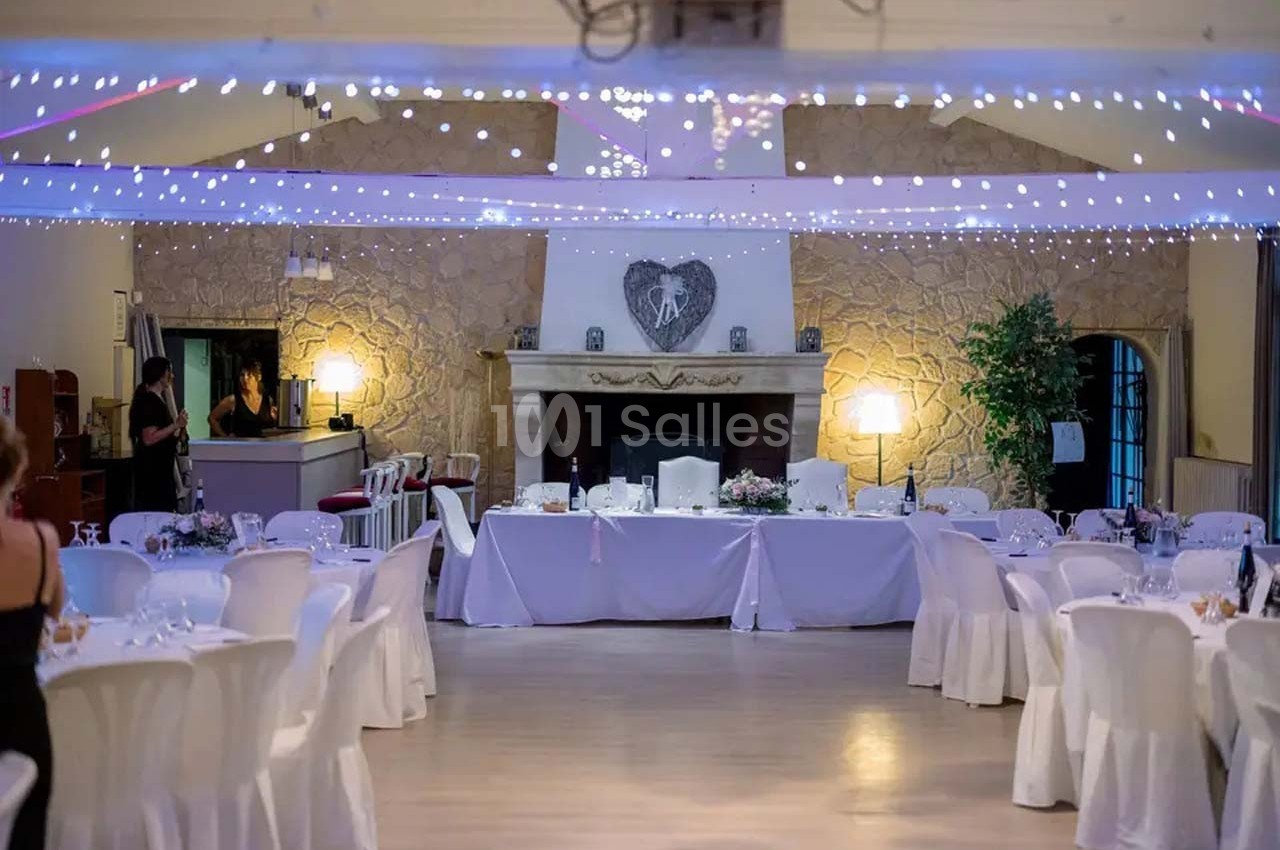 Salle de réception décorée avec des tables rondes, nappes blanches, guirlandes lumineuses et une cheminée en pierre.