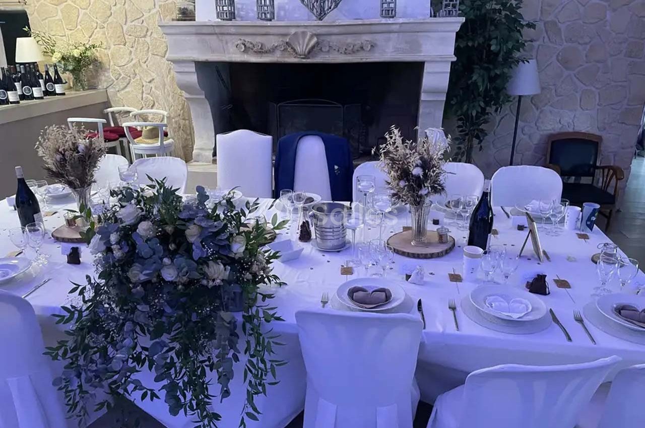 Table élégamment dressée pour un repas, décorée de fleurs séchées et entourée de chaises blanches, devant une cheminée en…