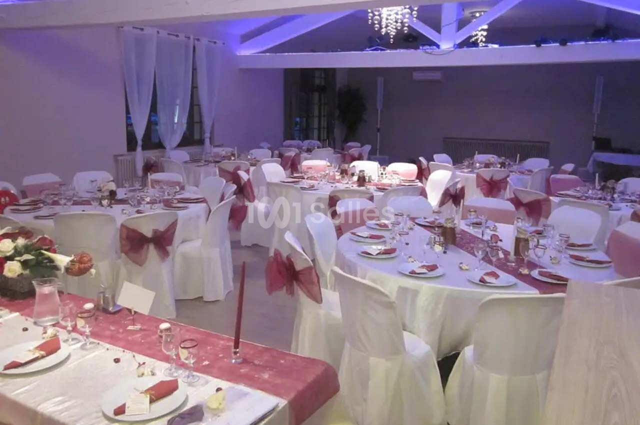 Salle de réception décorée avec des tables rondes, nappes blanches, chemins de table roses et chaises assorties.