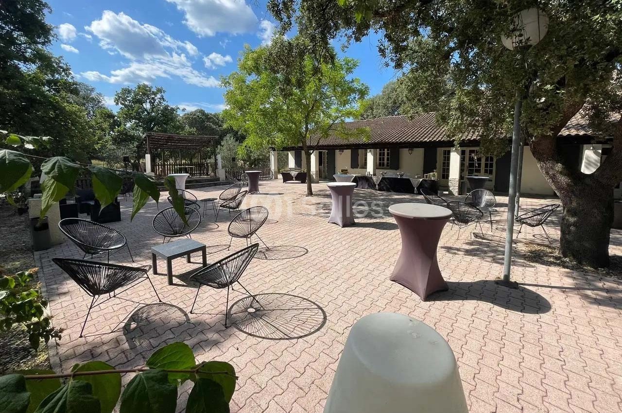 Terrasse pavée avec tables hautes, chaises design et arbres, devant un bâtiment à toit en tuiles sous un ciel dégagé.